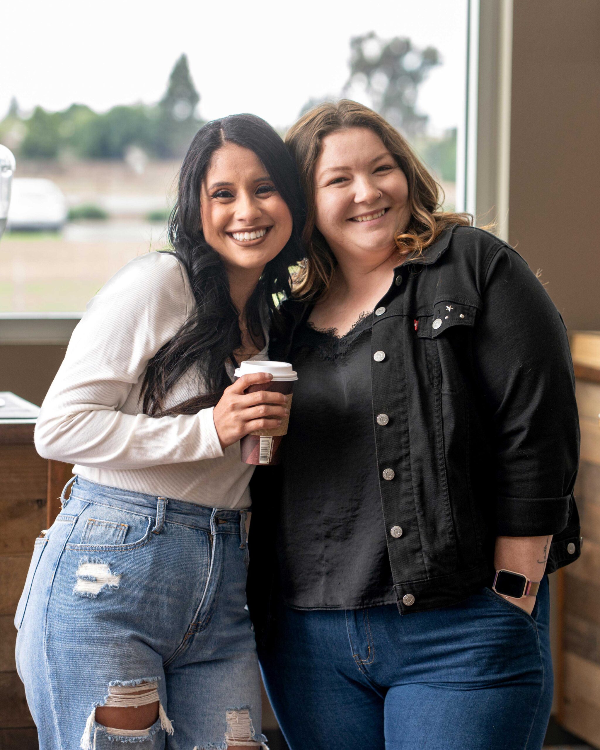 Two women smiling together with their arms around each other, holding coffee