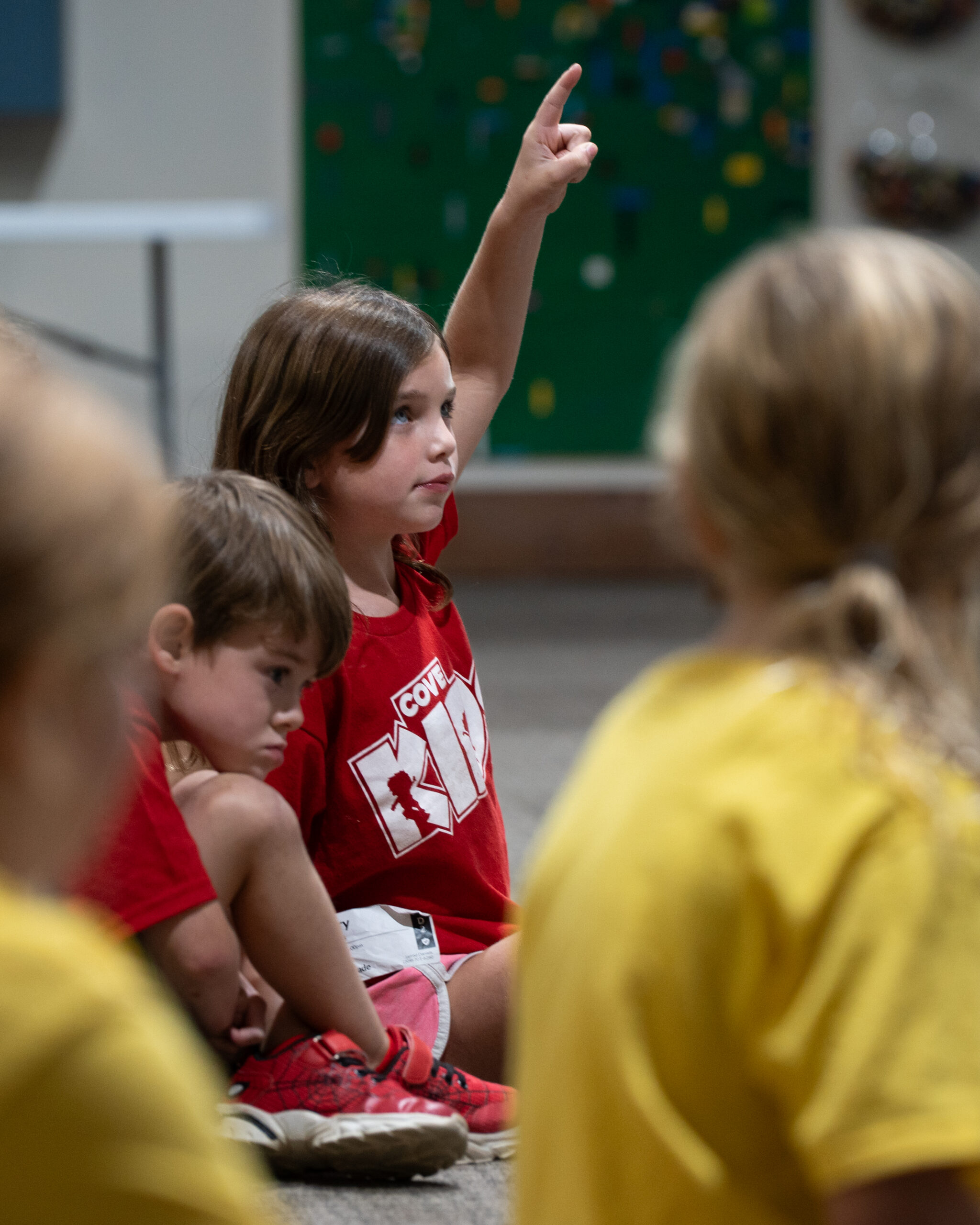A child raising their hand in a Cove Kids classroom with a climbing wall in the background