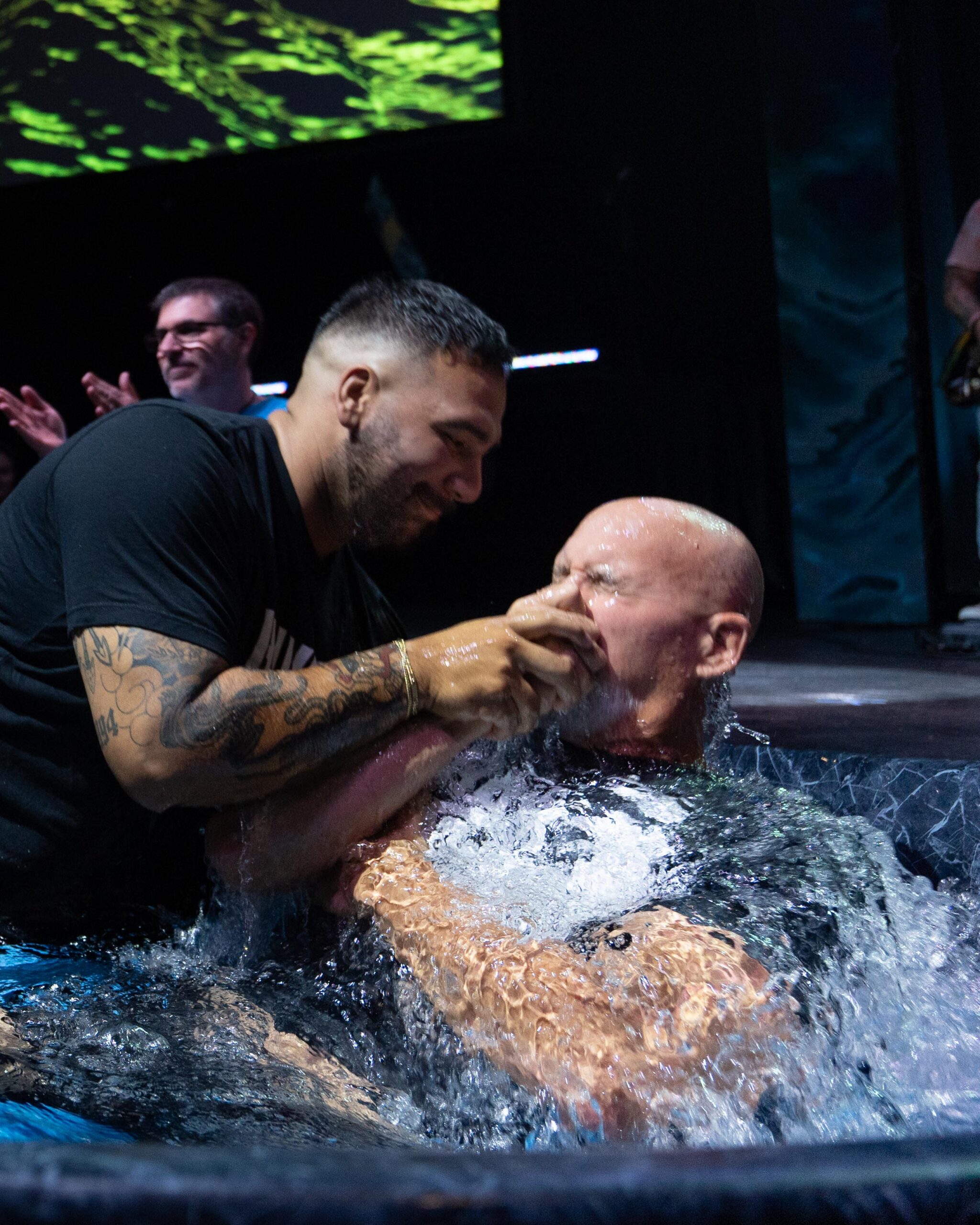 A bald man being baptized and held by a Shelter Cove pastor, water splashing during a Sunday baptism service