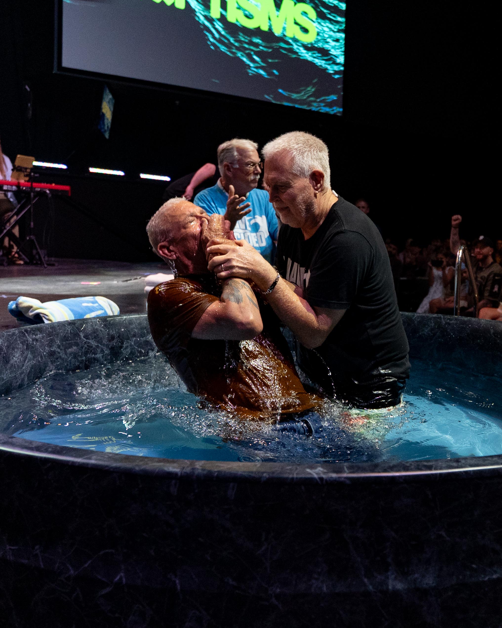 A man is baptized in the baptism tub on stage as another man holds him, with BAPTISMS displayed on the screen behind them at Shelter Cove Community Church
