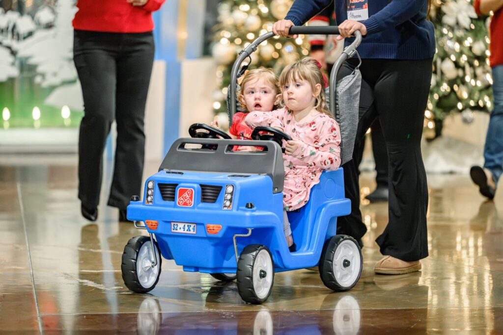 Two toddlers riding in a blue push car through the church lobby